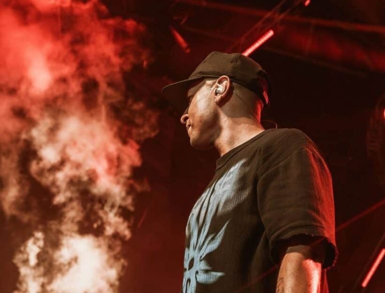 A man in a black t-shirt sits on a rooftop at sunset, gazing seriously at a woman walking away, creating a mood of reflection and emotional tension. LandonBuford.com