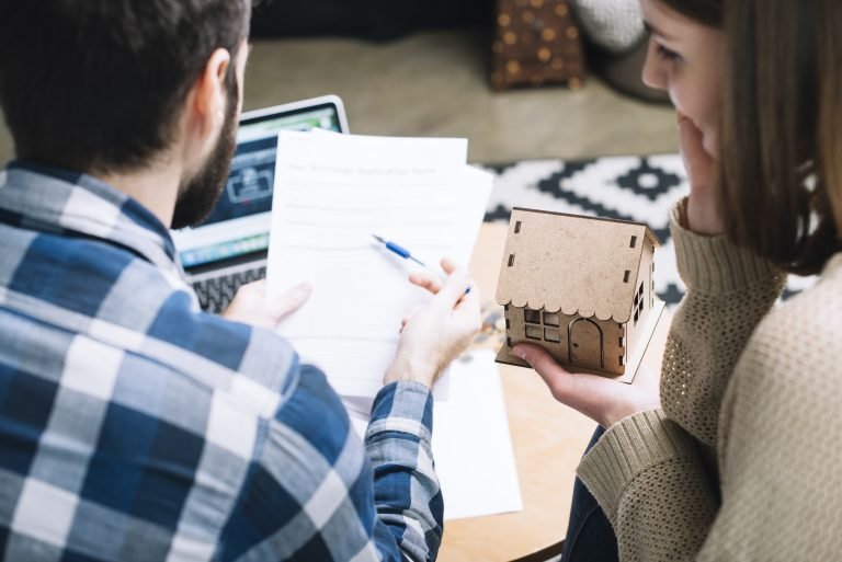 A couple reviews a mortgage application form together at a table, with a laptop in the background. One person is holding a pen and the form, while the other holds a small model house. LandonBuford.com