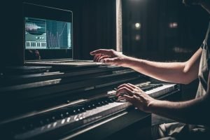 A person playing a digital piano while using a laptop displaying audio editing software, symbolizing the fusion of music production and digital distribution. LandonBuford.com
