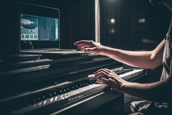 A person playing a digital piano while using a laptop displaying audio editing software, symbolizing the fusion of music production and digital distribution. LandonBuford.com