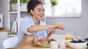 Woman sitting at a kitchen table enjoying a bowl of fresh fruits, promoting healthy and organic eating habits. LandonBuford.com