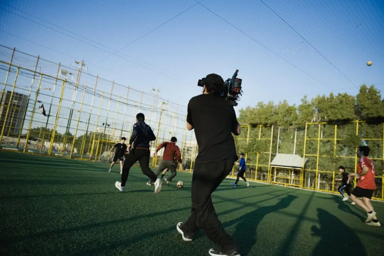 A camera operator films a group of young men playing soccer on an outdoor turf field enclosed by a tall yellow fence, capturing action during a bright afternoon. LandonBuford.com
