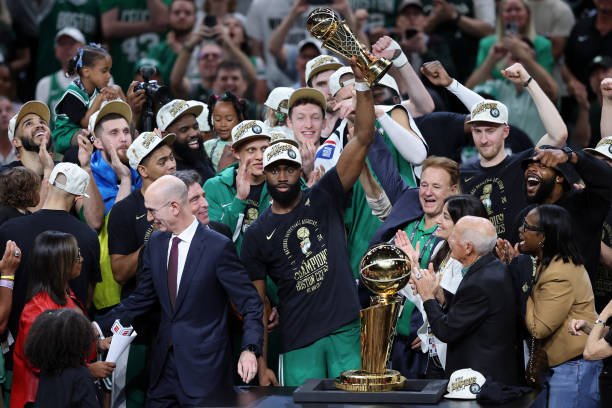 Jaylen Brown holds up the NBA Finals MVP trophy while surrounded by teammates, coaches, and fans celebrating the Boston Celtics' championship victory. LandonBuford