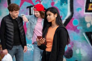 Teenage girl wearing wireless earbuds and holding a phone stands confidently in front of a colorful graffiti wall, while two other teens chat and smile in the background. LandonBuford