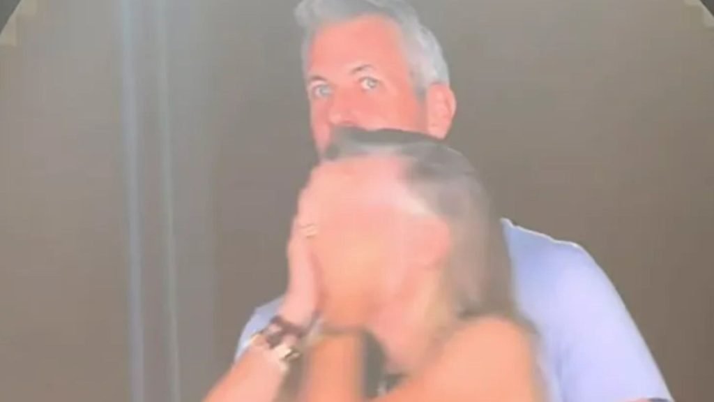 Astronomer CEO Andy Byron with gray hair looking directly into the camera while standing behind Kristin Cabot who is covering her face with her hands, both caught under bright stadium lighting.