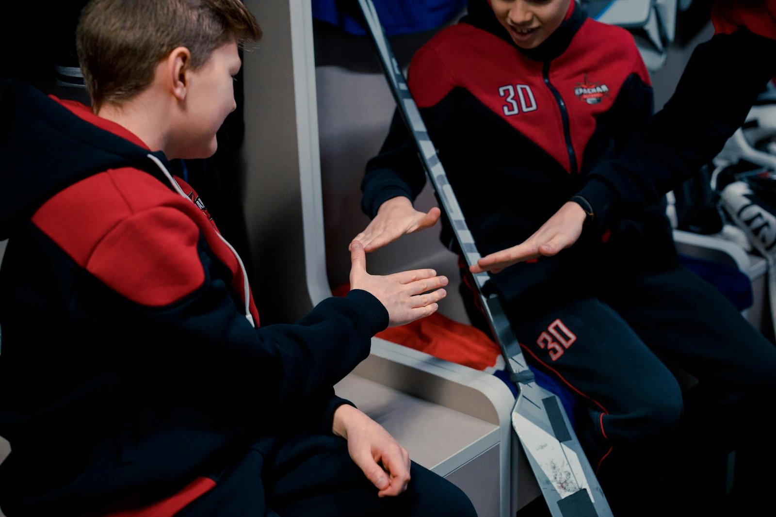 Two young boys wearing matching red and black tracksuits play a hand-slapping game while sitting in a locker room, smiling and enjoying their time together. LandonBuford.com