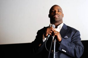 Former NFL player Ricky Williams speaks into a microphone while wearing a dark suit jacket and white shirt during a public appearance. LandonBuford.com