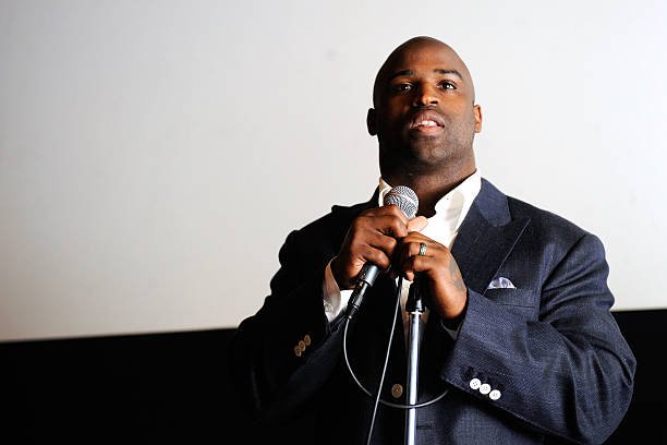 Former NFL player Ricky Williams speaks into a microphone while wearing a dark suit jacket and white shirt during a public appearance. LandonBuford.com